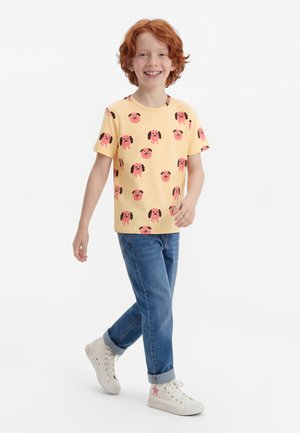 Κόκκινομαλλο παιδί που χαμογελά και περπατά, φορώντας κίτρινη μπλούζα με ροζ πρόσωπα σκύλων, μπλε τζιν και λευκά αθλητικά παπούτσια με ροζ αστέρι.