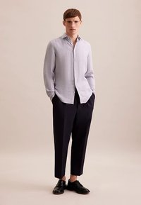 Camicia di lino azzurro chiaro, abbottonata sul davanti, abbinata a pantaloni scuri. Modello in piedi con le mani nelle tasche contro uno sfondo neutro.