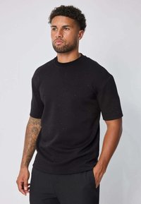 T-shirt noir à manches courtes avec un col rond, fabriqué en tissu lisse présentant des motifs discrets à pois. Le modèle se tient devant un fond neutre.