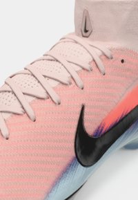 Nike futbola botas gaiši rozā krāsā ar strukturētu augšdaļu, melnu Swoosh logotipu un gradienta zila akcentiem tuvumā pie zoles. Redzamas sešas auklu caurumi.