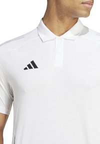 Weißes Poloshirt mit drei schwarzen Streifen auf der Schulter und einem schwarzen Adidas-Logo auf der Brust. Verfügt über einen klassischen Kragen und eine Knopfleiste.