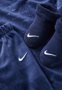Temno modro flis oblačilo, ki vključuje ujemajoče se hlače in copate z belimi Nike logotipi. Tkanina ima mehak občutek z rahlimi vzorci.