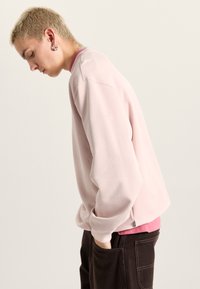 Lyserød sweatshirt med lange ærmer og rund halsudskæring. Har et kortere indre lag i en mørkere nuance, sideslids og manchetter ved ærmerne.