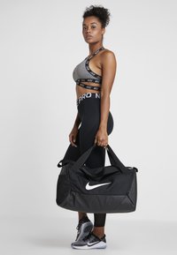 Czarna torba sportowa Nike z wyraźnym białym logo, w połączeniu z szarym biustonoszem sportowym i czarnymi legginsami. Teksturowany materiał i elegancki design.