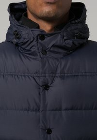 Mann, der eine navyblaue gepolsterte Jacke mit hohem Kragen und schwarzen Druckknöpfen trägt, sichtbar von Kinn bis Brust.