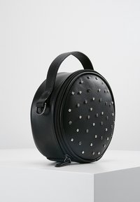 Rund sort faux lædertaske med sølvnitter, lynlåslukning og et hanke, der har en glat overflade og kompakt design.
