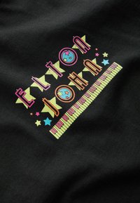 Schwarzer Stoff mit buntem grafischen Text "ELTON JOHN" sowie musikalischen Noten und einem Klavierdesign in den Farben Pink, Grün, Blau und Gelb.