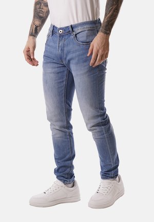 Uomo che indossa jeans aderenti azzurri chiaro, sneakers bianche e una camicia bianca, mostrando le braccia tatuate su uno sfondo semplice.