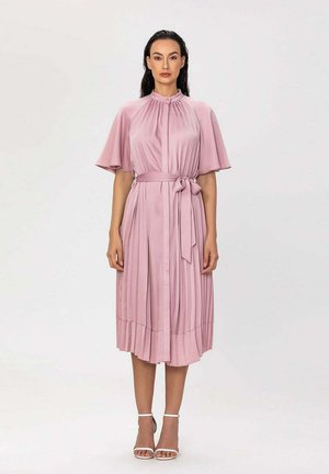 Femme portant une robe plissée rose clair longueur genou, manches courtes et ceinture à la taille, debout sur un fond blanc uni.
