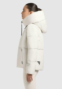 Giacca a vento bianca con collo alto e cappuccio oversize. Caratterizzata da tasche con zip e dettagli in nero sul manica. Texture liscia.