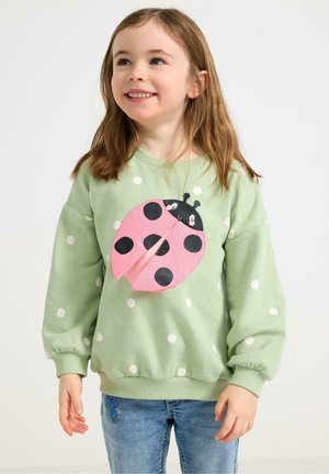 Young girl wearing light green polka dot sweatshirt with 3D pink and black ladybug design, paired with blue denim jeans.