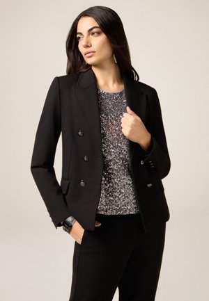 Blazer negro de doble botonadura con botones plateados, usado sobre una blusa de lentejuelas. El traje cuenta con un diseño ajustado y detalles a medida.