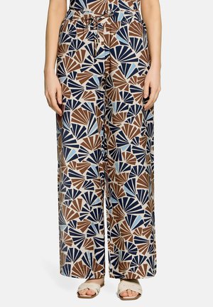 Weit geschnittene Hosen mit braunen, marineblauen und hellblauen fächerförmigen geometrischen Mustern, getragen mit weißen Sandalen von einer stehenden Person.