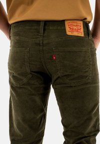 Pantalones de pana color verde oliva con bolsillos traseros, que cuentan con una etiqueta de marca de cuero marrón y una pequeña etiqueta roja en la parte trasera de la cintura.