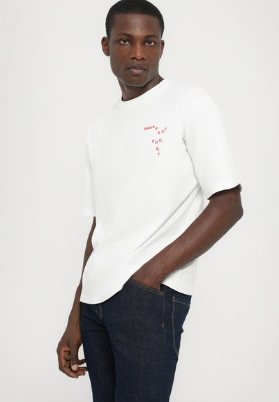 White short-sleeve t-shirt with a red graphic print on the chest; paired with dark blue jeans. Smooth fabric, relaxed fit.