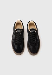 Zapatillas de ante y fieltro negras con un diseño de corte bajo, que cuentan con cordones negros, interior beige y una suela de goma texturizada.