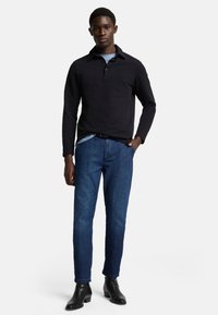 Polo a maniche lunghe nera indossata sopra una camicia azzurra chiara, abbinata a jeans denim blu scuro e stivali neri alla caviglia. Design semplice e vestibilità rilassata.
