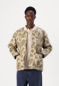 Polstret bomberjakke i et beige camouflage-mønster, med trykknaplukning fortil, sidelommer og et logo-patch på brystet.
