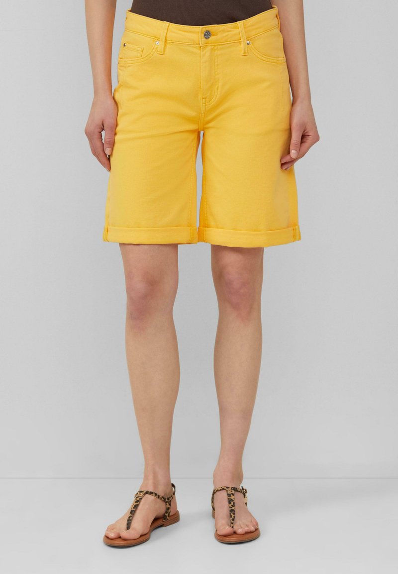 Shorts en denim jaunes avec ourlets retroussés, dotés de poches avant et d'une fermeture à bouton en métal. Associés à des sandales à imprimé animal.