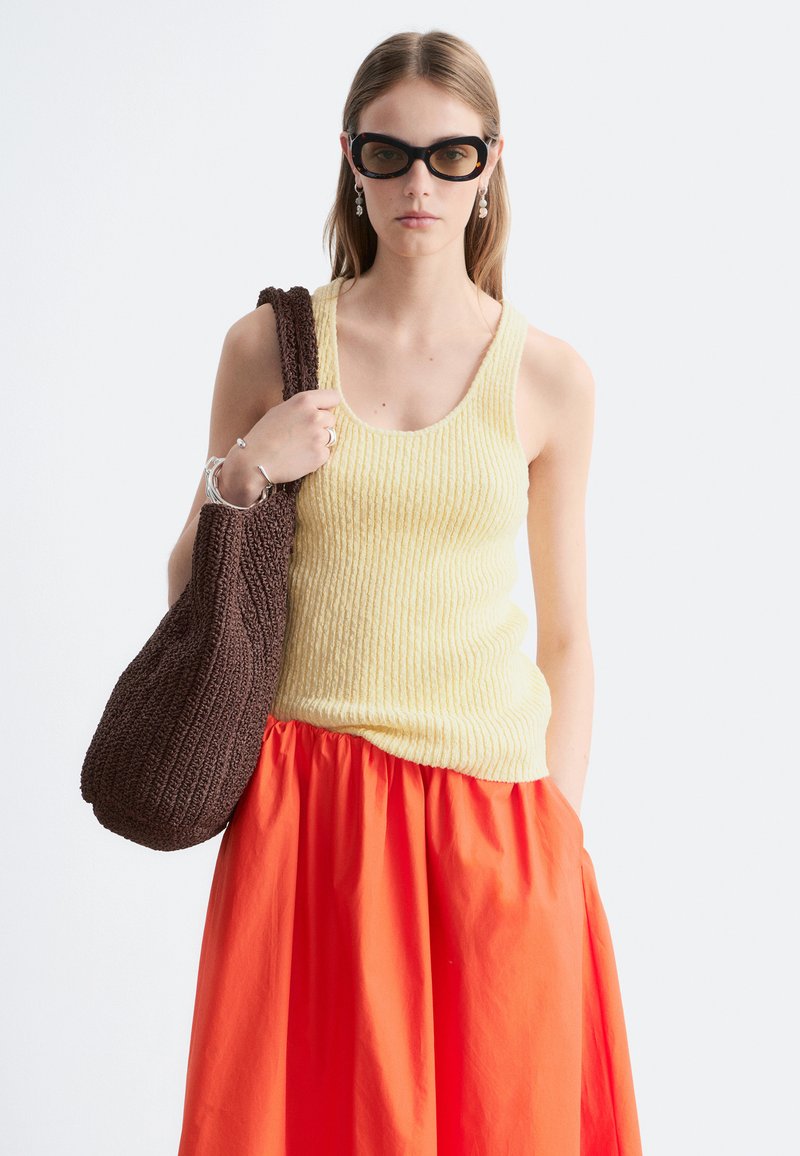 Femme portant un haut jaune sans manches, jupe orange, lunettes de soleil noires, tenant un grand sac cabas marron tricoté sur l'épaule.