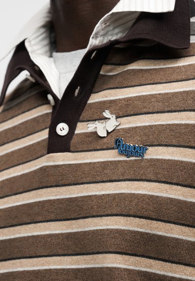 Καφέ ριγωτό πόλο πουκάμισο με σκούρο γιακά, που διαθέτει ένα ασημί σμάλτο καρφίτσα με δύο κουνέλια και ένα μπλε λογότυπο με την επιγραφή "Amour Toujours".