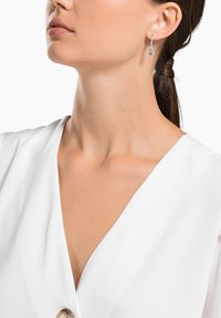 Silver earrings with dangling design featuring small blue crystals, showcasing a minimalist aesthetic. Model wears a white V-neck top.