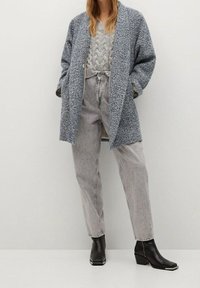 Cappotto azzurro chiaro a spina di pesce sopra un maglione grigio a trecce, abbinato a jeans grigio chiaro e stivaletti neri con punta quadrata e dettagli.