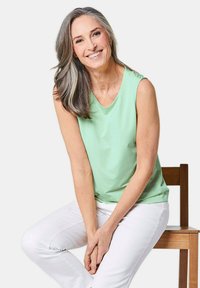 Lichtgroene mouwloze top met een ronde hals, gecombineerd met een witte broek. Glad materiaal met een losse pasvorm. Model zit op een houten stoel.