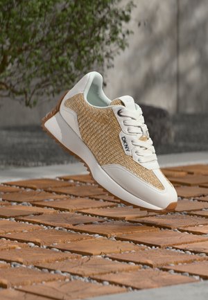 Witte en beige geweven sneaker met gumzool en DKNY-logo, zwevend boven een getextureerd bruin bakstenen trottoir buiten.