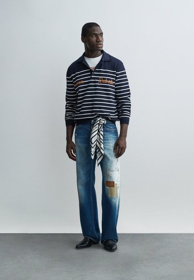 Homme debout portant une chemise à manches longues rayée bleu marine avec le texte « Rebels », un jean usé patchwork, une ceinture écharpe à motifs, et des chaussures noires.