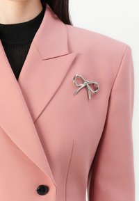 Růžový blazer s klopovým límcem, doplněný černým žebrovaným topem pod ním. Na klopě je výrazně umístěna brož ve tvaru stříbrné mašle.