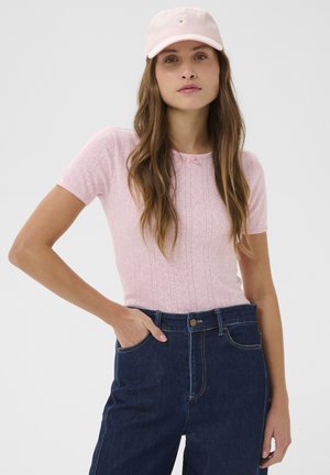 Femeie purtând un tricou roz cu mâneci scurte din tricot, blugi înalți pe talie, de culoare albastru închis, și o șapcă roz deschis cu broderie albă de margaretă.