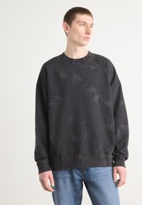 Sudadera negra con un patrón ligeramente desgastado, cuello redondo y mangas largas. Tejido suave y texturizado; combinada con jeans de un azul claro.