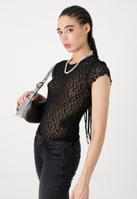 Schwarzes Spitzenoberteil mit kurzen Ärmeln, figurbetontem Design und gewellten Rändern. Kombiniert mit schwarzen Jeans und einer silbernen Handtasche mit strukturiertem Finish.