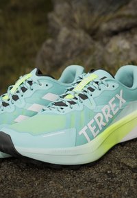Svetlomodré trailové bežecké topánky Adidas Terrex s čiernymi podrážkami a žltými akcentmi na štrku vonku.
