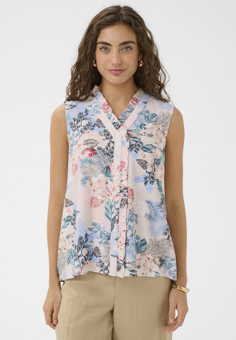 Femme aux cheveux bouclés portant un chemisier sans manches à boutons avec motif floral pastel et un pantalon beige, debout devant un fond blanc uni.