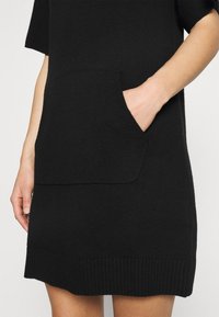 Robe noire en tricot avec manches courtes, dotée d'une grande poche frontale et d'un ourlet côtelé. Texture douce et coupe décontractée.
