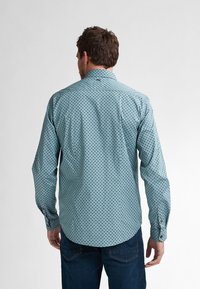 Camicia a maniche lunghe di colore azzurro con un motivo scuro ripetitivo, colletto, polsini con bottoni e orlo curvo, indossata con jeans scuri.