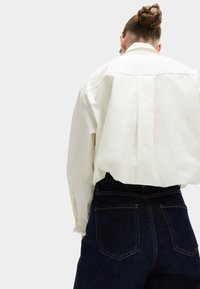 Camicia bianca oversize con maniche lunghe, caratterizzata da una piega sul retro, abbinata a jeans in denim scuro a vita alta con due tasche posteriori.