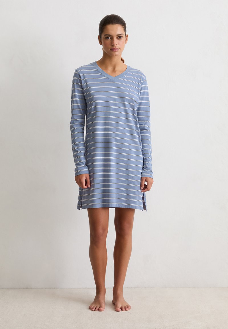 Mujer de pie descalza, vistiendo una camisa de dormir de manga larga a rayas horizontales azul y blanca con aberturas laterales, contra un fondo blanco liso.