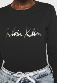 Svart långärmad t-shirt i bomull med en silverreflekterande "Calvin Klein" logotyp tryckt på framsidan, som kombineras med denim shorts.