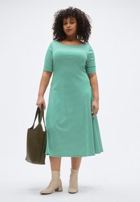 Zöld-fehér csíkos ruha rövid ujjakkal, bővülő szoknyával, bézs bokacsizmával és sötétzöld táskával viselve.