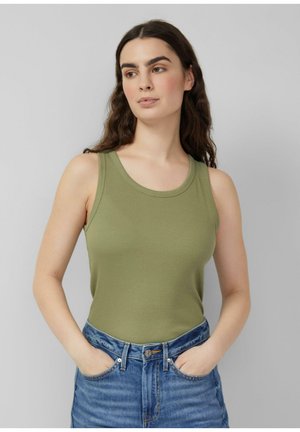 Młoda kobieta o długich ciemnych włosach, ubrana w oliwkowo-zielony top na ramiączkach i niebieskie dżinsy, stojąca z rękami w kieszeniach i patrząca na bok.