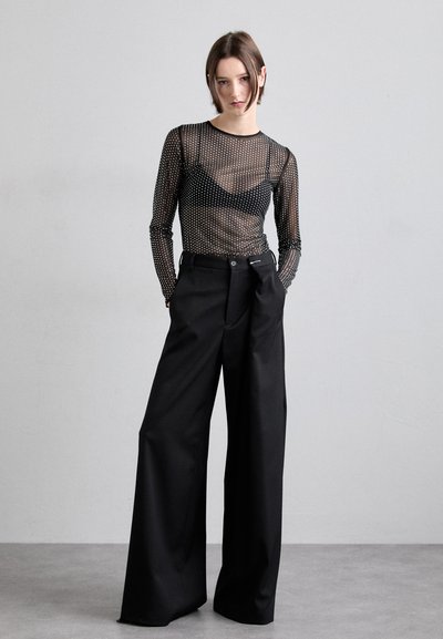 Schwarzes Mesh-Oberteil mit langen Ärmeln und weißen Punkten, das über einem schwarzen Bralette getragen wird, kombiniert mit hochgeschnittenen, locker sitzenden schwarzen Hosen.