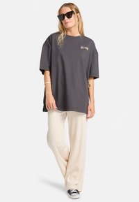 Žena v oversized tmavě šedém tričku, krémových širokých kalhotách s ribbováním, černých teniskách a velkých černých slunečních brýlích, stojící s jednou nohou zkříženou.