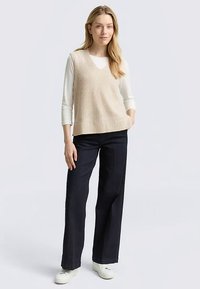 Gilet lavorato a maglia beige con scollo a V, indossato sopra una camicia bianca a maniche lunghe. Abbinato a jeans scuri a gamba larga e sneakers bianche. Tessuto liscio.