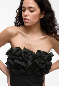 Czarny top bez ramiączek z falbaniastym dekoltem. Gładka góra z żebrowanym wzorem, eksponująca warstwowe, obszerne fałdy materiału.