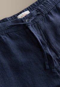 Pantalon en lin bleu marine avec taille élastique, bouton et cordon de serrage noué, affichant une étiquette "Next Signature" à l'intérieur de la ceinture.