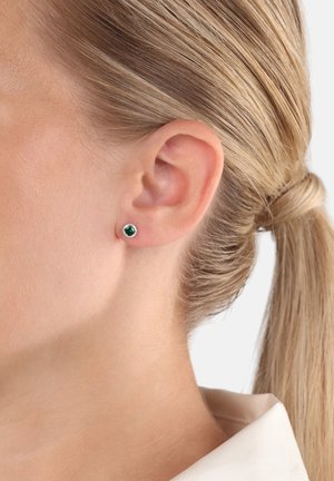 Primo piano di un orecchio femminile con capelli biondi raccolti, che indossa un piccolo orecchino a bottone d'argento con una gemma verde scuro.