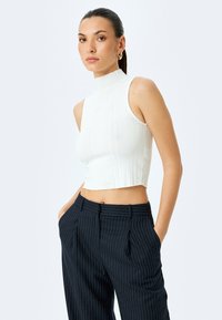 White sleeveless ribbed crop top with a high neckline, paired with navy pinstriped trousers with side pockets and pleated front details.